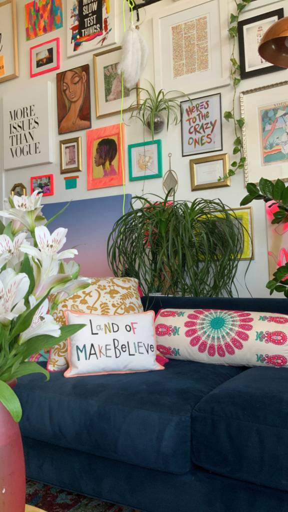 Un salon coloré au décor maximaliste, comprenant un canapé bleu avec des coussins décoratifs, entouré d'œuvres d'art et de plantes.