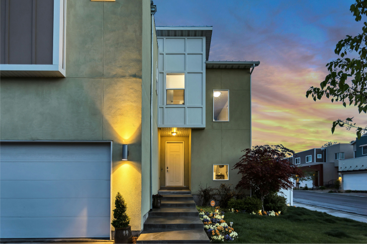 Maison moderne de deux étages avec extérieur beige et garage attenant, éclairée au crépuscule sous un ciel coucher de soleil coloré.