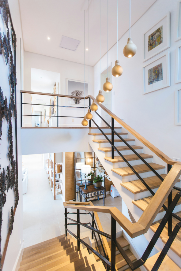 Intérieur de maison moderne comprenant un escalier en bois avec des balustrades en métal noir menant à un loft, avec des suspensions et un couloir blanc décoré d'œuvres d'art.