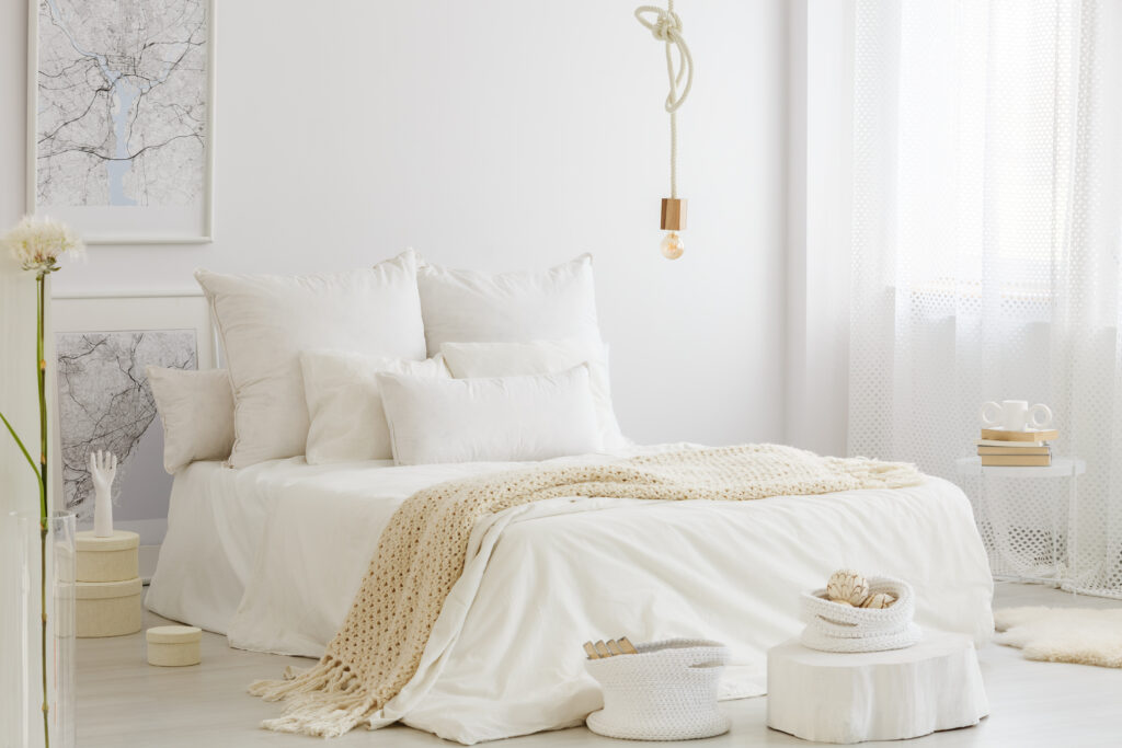 Une chambre minimaliste avec un lit blanc, plusieurs oreillers, une couverture en tricot beige et des objets de décoration blancs. Une carte encadrée, une ampoule suspendue et des rideaux blancs transparents complètent la pièce.