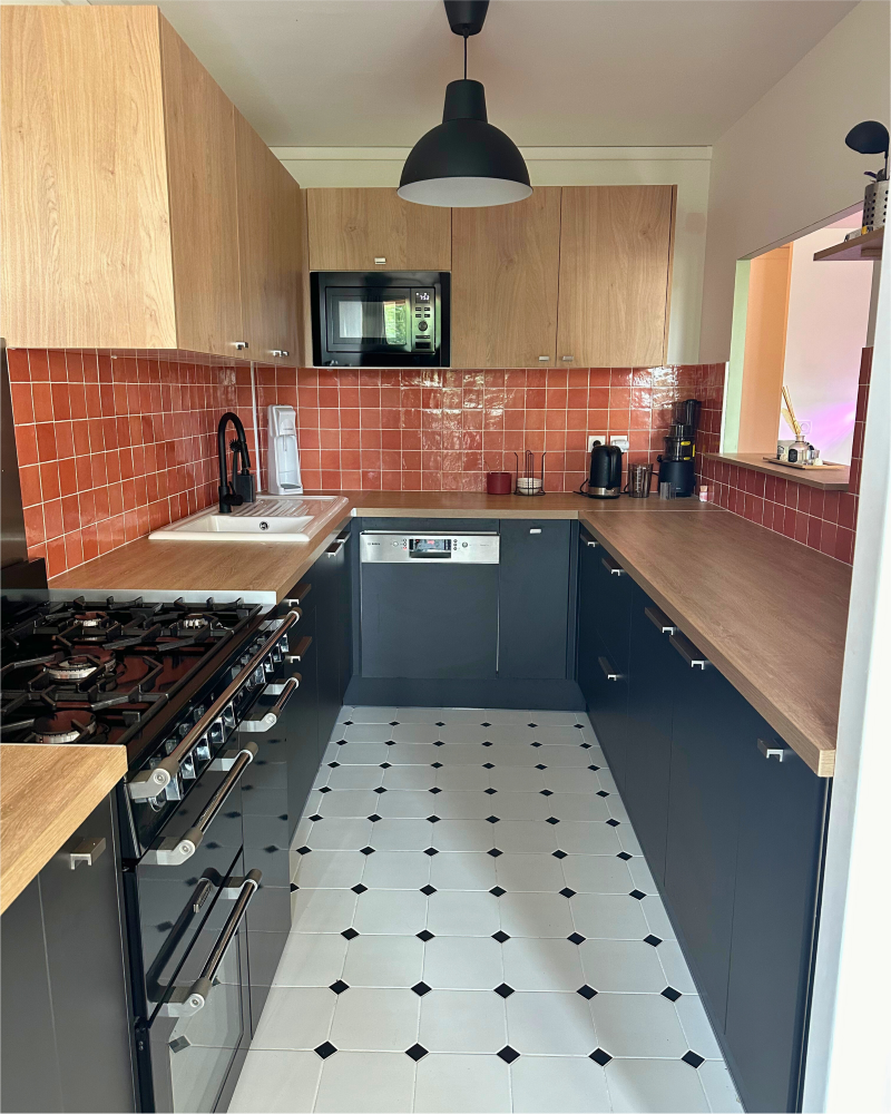 Cuisine moderne avec armoires en bois et gris foncé, dosseret en carreaux rouges, comptoirs blancs, électroménagers noirs, plancher blanc avec des accents noirs et luminaire suspendu noir — cuisine rénovée par Originatelier.