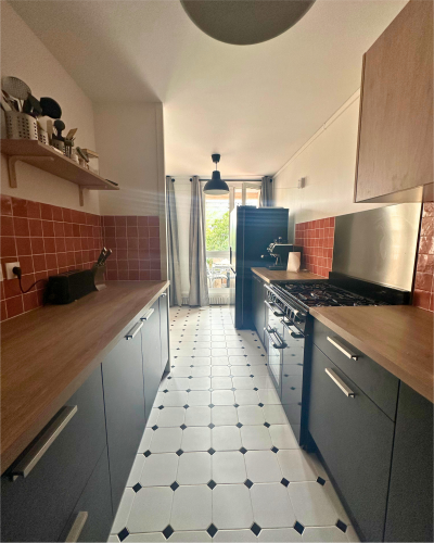 Cuisine étroite avec comptoirs en bois, dosseret en carrelage rouge, armoires noires et sol en carrelage blanc. Une fenêtre au fond apporte de la lumière naturelle. Un réfrigérateur et une cuisinière se trouvent sur les côtés opposés de cette cuisine rénovée par Originatelier.
