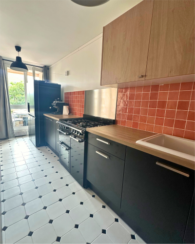 Une cuisine étroite avec des armoires noires et en bois, un dosseret en carrelage rouge et une cuisinière autoportante. Une fenêtre au bout laisse entrer la lumière naturelle. Le sol est carrelé blanc avec un motif de losanges noirs, le tout faisant partie d'une superbe cuisine rénovée par Originatelier.