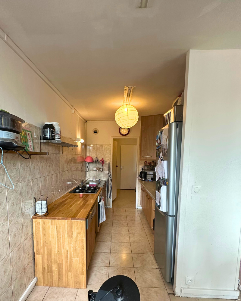 Une cuisine étroite aux murs carrelés est dotée de comptoirs en bois et de divers appareils électroménagers. Des luminaires suspendus illuminent l'espace. Un réfrigérateur trône sur le côté droit près de l'entrée, mettant en valeur une cuisine à rénover confiée à Originatelier.