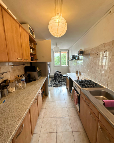 Une cuisine étroite avec des armoires en bois, des comptoirs en granit, une cuisinière à gaz, un évier et des sols carrelés. Le mur du fond est doté d'une fenêtre et d'un coin repas avec une table et des chaises, un espace charmant pour une cuisine à rénover confiée à Originatelier.