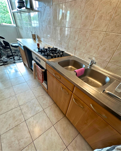 Une cuisine aux murs carrelés, un évier avec une éponge rose, une cuisinière à gaz, des armoires en bois et un comptoir avec des ustensiles de cuisine : une charmante cuisine à rénover confiée à Originatelier.