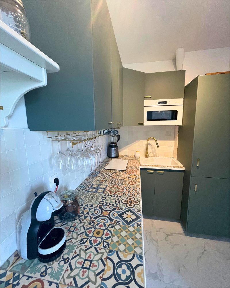 Une petite cuisine moderne avec des armoires vertes, un comptoir à motifs, un dosseret blanc, une machine à expresso, des verres à vin suspendus, une bouilloire électrique et un four à micro-ondes intégré; cette cuisine rénovée par Originatelier magnifiquement conçue respire à la fois le style et la fonctionnalité.