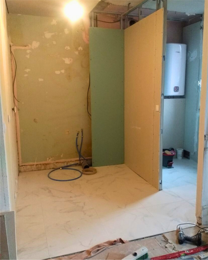 Une pièce partiellement construite avec des murs en cours de construction, quelques outils et matériaux sur le sol et une zone inachevée à l'arrière avec un chauffe-eau. Cette cuisine confiée à Originatelier pour rénovation est déjà prometteuse avec son travail artisanal détaillé.