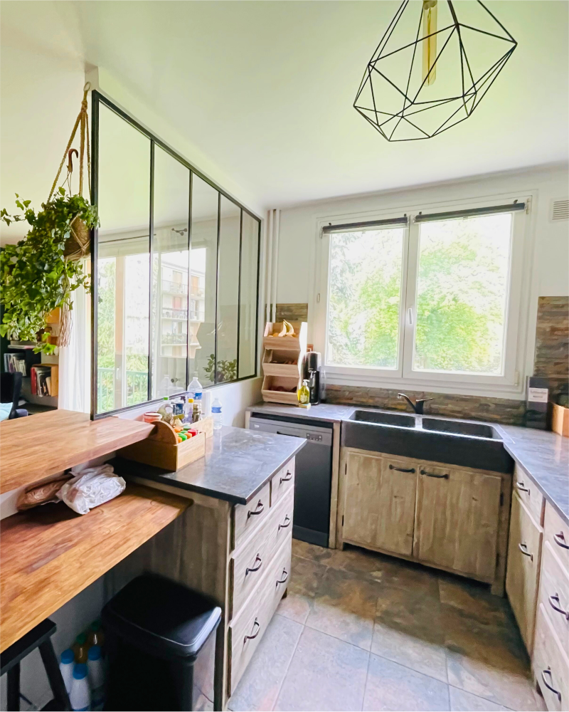 Une cuisine bien éclairée avec des armoires en bois, une grande fenêtre, une suspension géométrique et une plante suspendue reflète l'art de la cuisine rénovée par Originatelier.