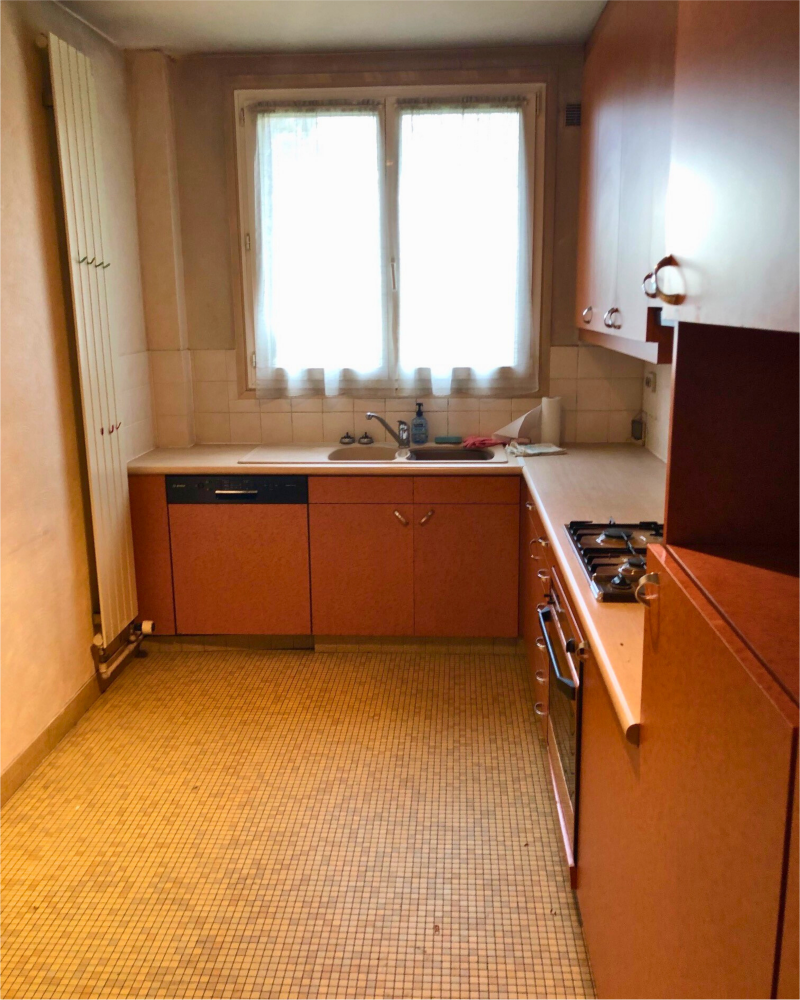 Une cuisine pittoresque avec des armoires orange, une cuisinière à gaz, un évier et un radiateur attend sa touche de transformation. Le sol est carrelé de beige, tandis qu'un rideau transparent orne la fenêtre.