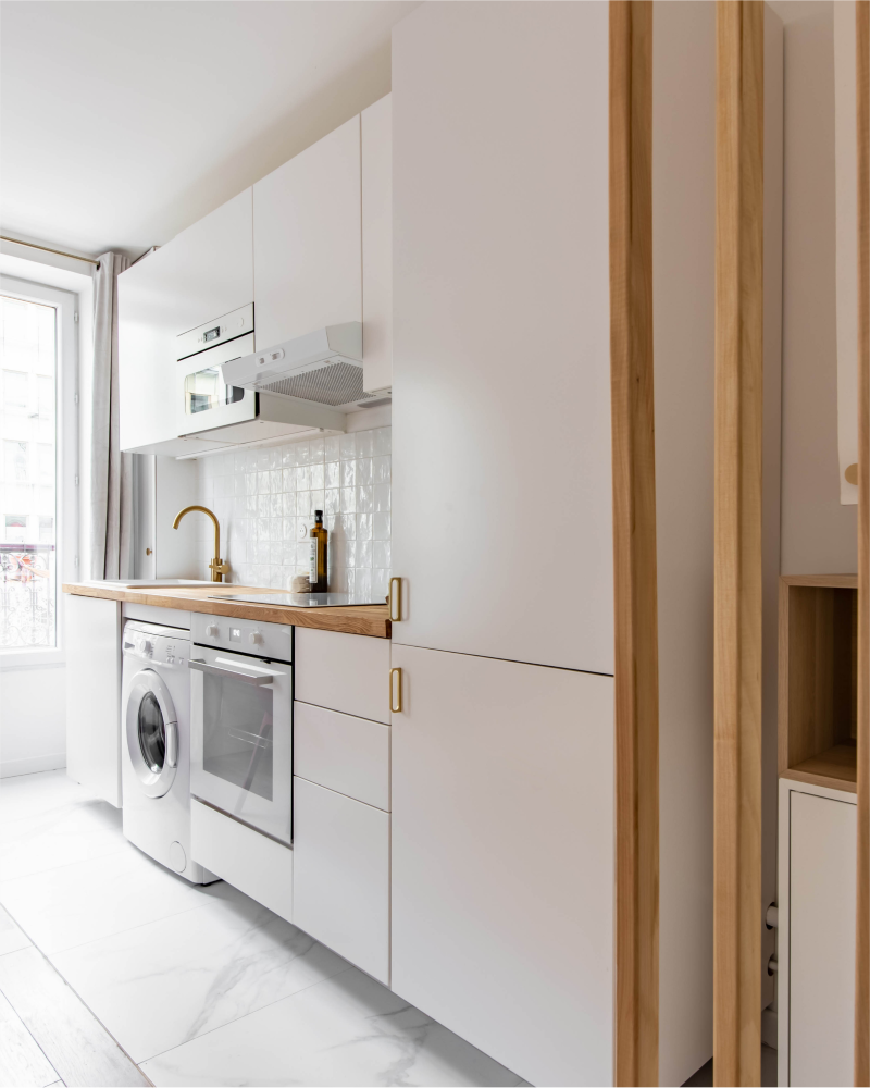 Une cuisine compacte et moderne signée Originatelier avec des armoires blanches, une machine à laver, un four, un comptoir en bois, un robinet doré et un dosseret carrelé. Parfaitement positionné à côté d'une fenêtre avec des rideaux, cet espace attend que votre touche personnelle brille.