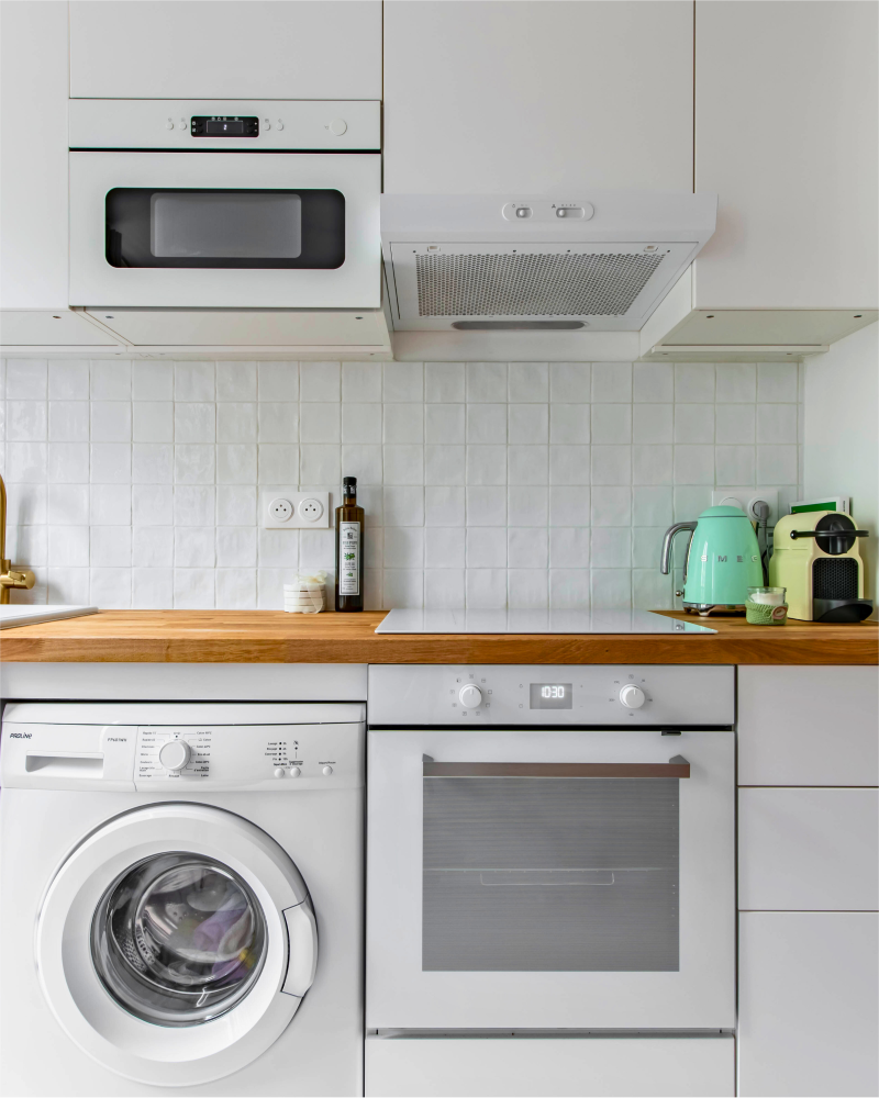 La cuisine moderne, rénovée par Originatelier, comprend un lave-linge, un four, un micro-ondes et une cafetière. Des armoires blanches et un dosseret carrelé complètent le design. Une bouilloire et une bouteille d'huile d'olive reposent gracieusement sur le comptoir en bois.