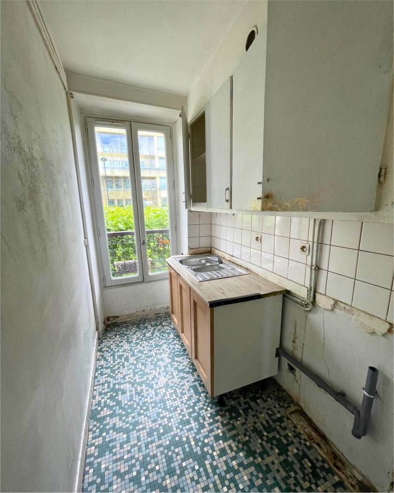 Cuisine étroite avec un petit évier, des armoires en bois et un dosseret carrelé. Le sol est orné d'un motif en mosaïque, tandis qu'une fenêtre laisse entrer la lumière naturelle. La peinture écaillée sur les murs indique qu'il s'agit d'une cuisine à rénover.