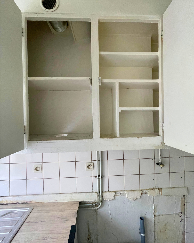 Des armoires de cuisine vides et usées avec de la peinture écaillée au-dessus d'un dosseret carrelé mettent en valeur cet espace, mettant en valeur son potentiel en tant que cuisines à rénover. Avec des portes d'armoires inférieures manquantes et une plomberie exposée sous la zone de l'évier, il y a de la place pour la créativité dans la revitalisation de cette cuisine.