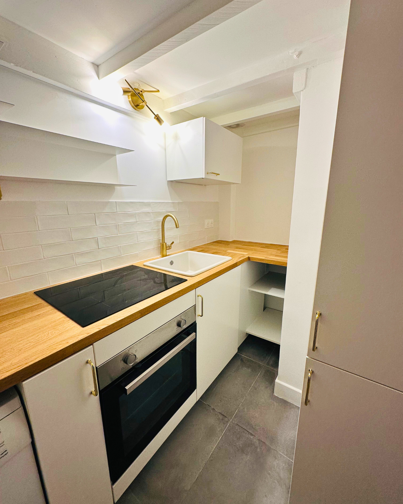 Une petite cuisine moderne avec un dosseret en carrelage blanc, des comptoirs en bois, une cuisinière électrique, un four, un évier blanc avec un robinet doré et des armoires blanches.