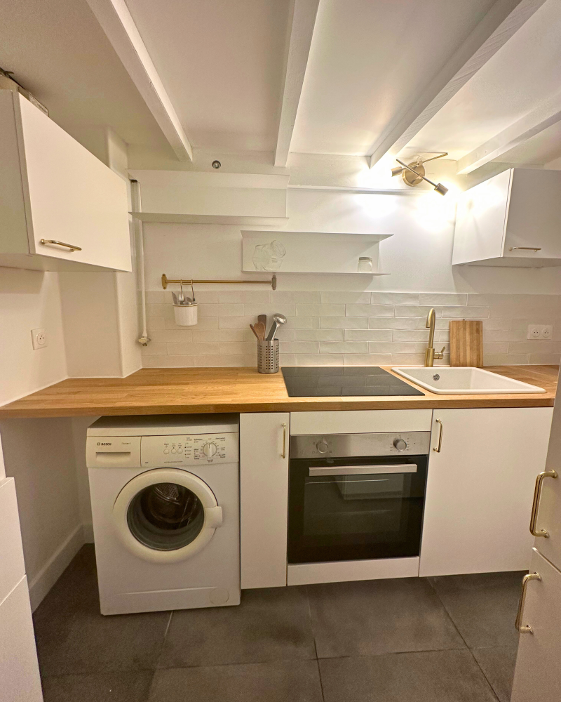 Une petite cuisine avec un micro-ondes sur le réfrigérateur, une bouilloire sur la machine à laver et une cuisinière à côté. Un carrelage décoratif avec un motif de fruits est sur le mur.