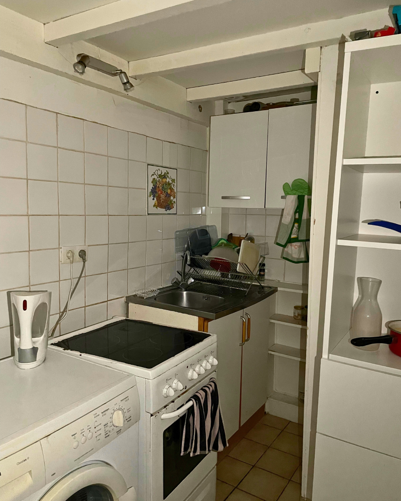 Cuisine_6_Avant_A Petite cuisine avec cuisinière blanche, machine à laver et placards. Un évier avec de la vaisselle qui sèche, un mur carrelé et une bouilloire sur le comptoir. Petite étagère avec divers articles de cuisine.