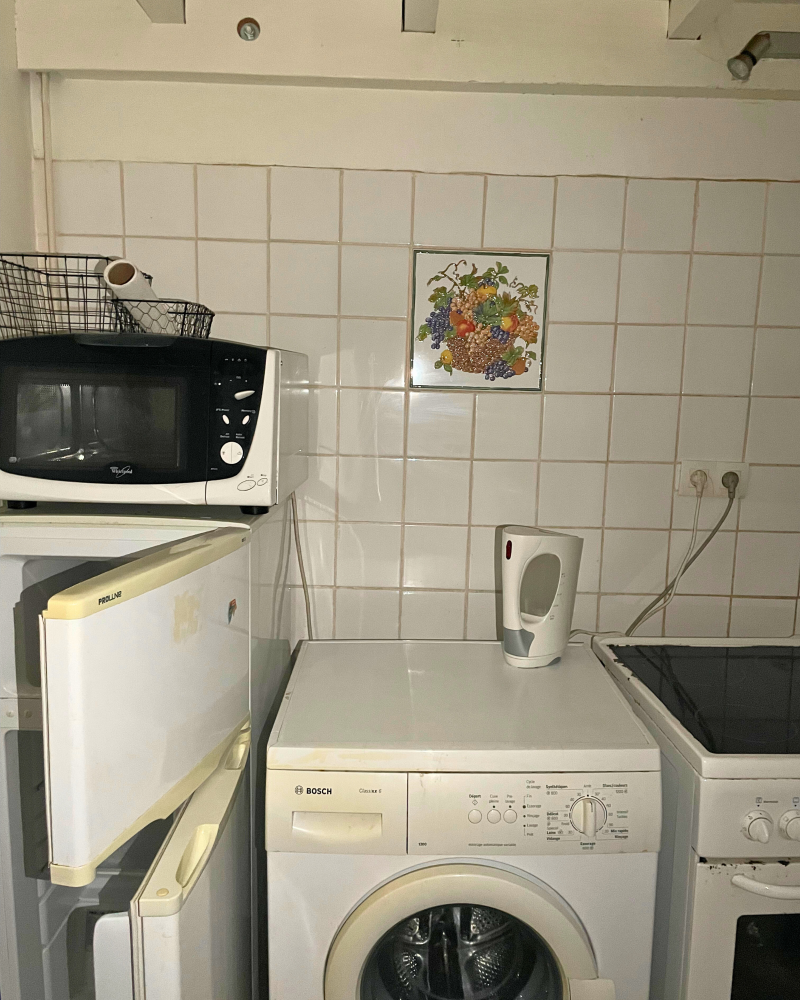 Cuisine_6_Avant_C Une petite cuisine avec un micro-ondes sur le réfrigérateur, une bouilloire sur la machine à laver et une cuisinière à côté. Un carrelage décoratif avec un motif de fruits est sur le mur.