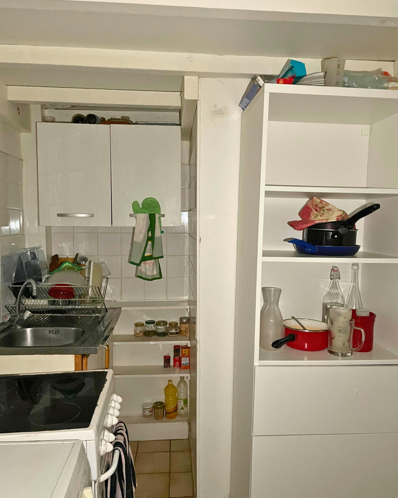 Cuisine_6_Avant_E Cuisine compacte avec un petit évier, un égouttoir, une cuisinière et des étagères contenant des casseroles, des bouteilles et des épices. Des armoires blanches offrent du rangement.