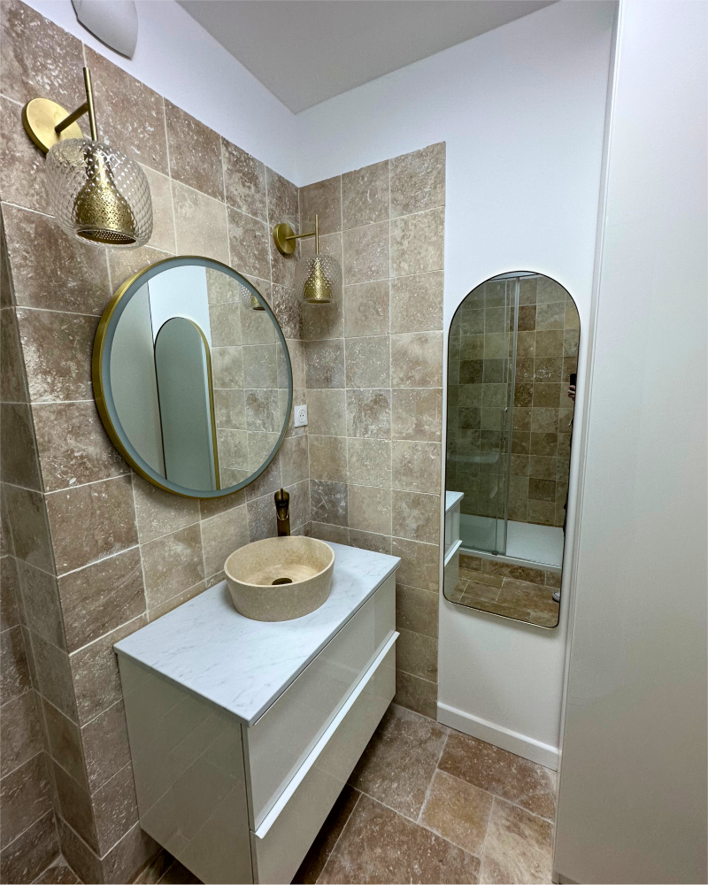 Salle de bain avec murs carrelés beiges, un miroir rond au-dessus d'une vanité blanche avec une vasque en pierre, une applique murale dorée et un miroir ovale à droite.