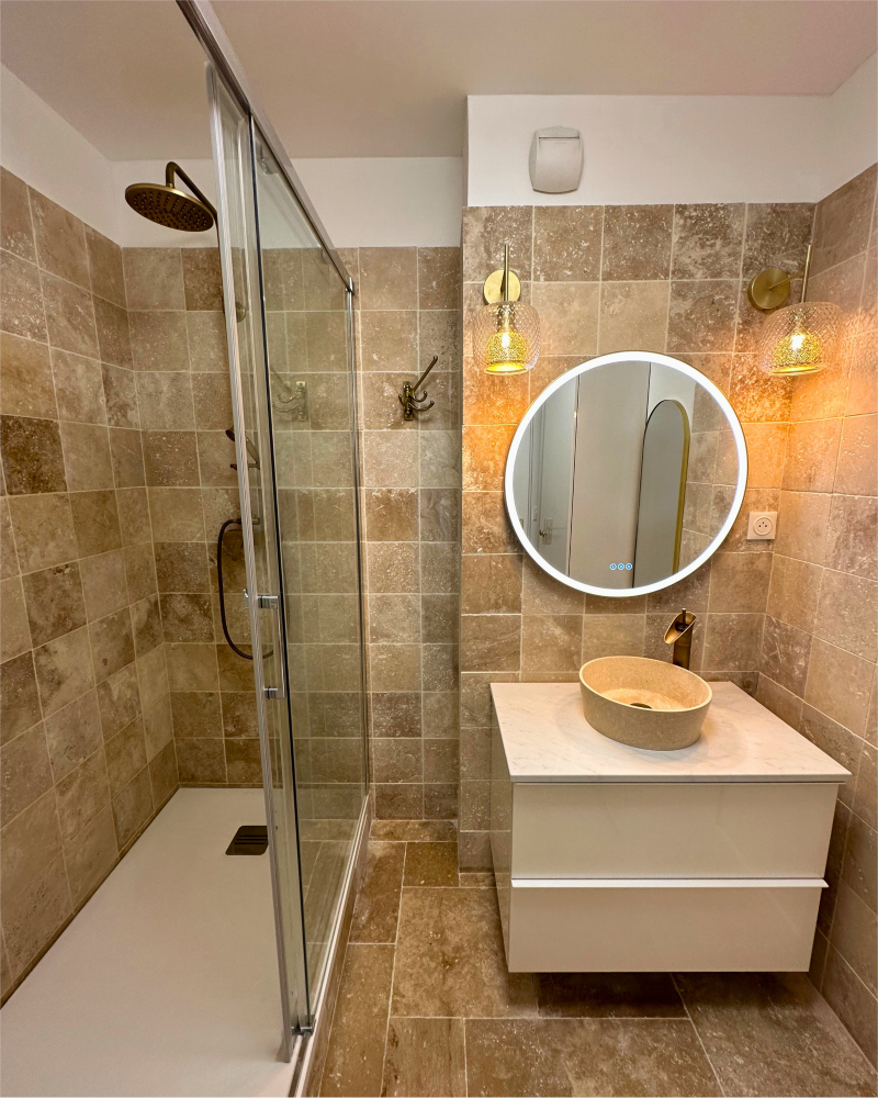 Salle de bain moderne avec douche et murs carrelés en pierre. Un miroir rond est suspendu au-dessus d'un lavabo minimaliste avec vasque en pierre. Deux suspensions sont placées de chaque côté du miroir.