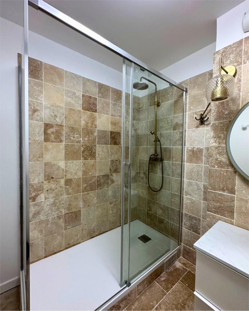 Une salle de bain moderne avec une douche vitrée avec des carreaux de pierre beige, une pomme de douche à effet pluie et un miroir rond mural au-dessus du lavabo.