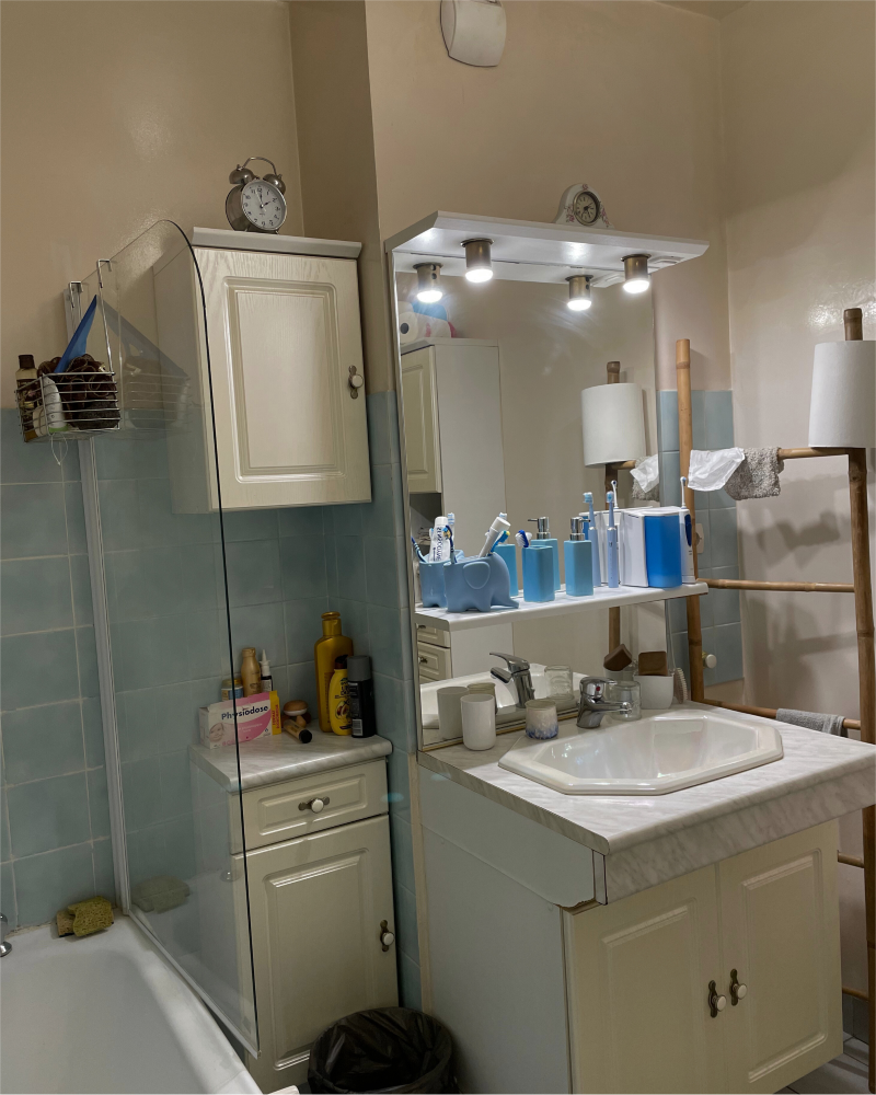 Une salle de bain avec un lavabo Une salle de bain avec un lavabo, un miroir et un meuble. Divers produits de toilette et des contenants bleus sont posés sur le comptoir. Une baignoire avec une cloison en verre et un porte-serviettes sont visibles.