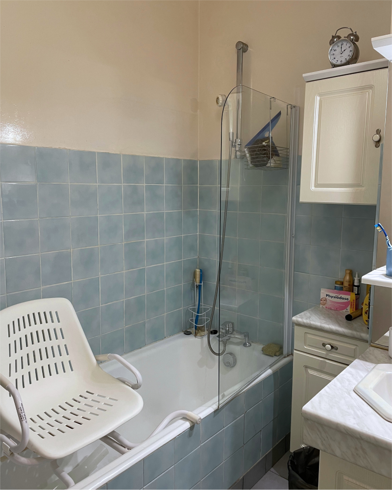 SDB_2_Avant_E Salle de bain avec murs carrelés bleus, baignoire avec chaise de douche blanche, paroi de douche en verre et une armoire avec des articles de toilette et une horloge sur le dessus.