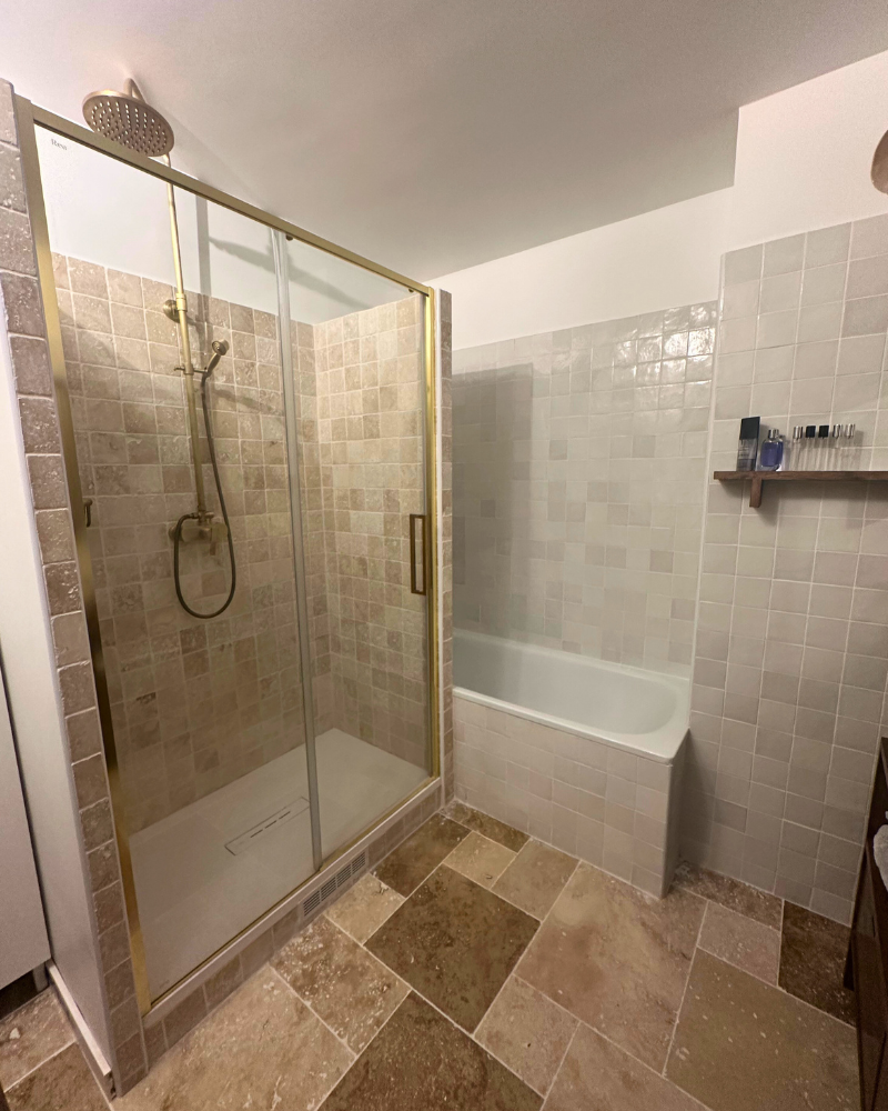 Salle de bain avec cabine de douche en verre, pomme de douche à effet pluie et baignoire adjacente. Des carreaux beiges et crème recouvrent les murs et le sol, avec une petite étagère pour ranger les produits de toilette.