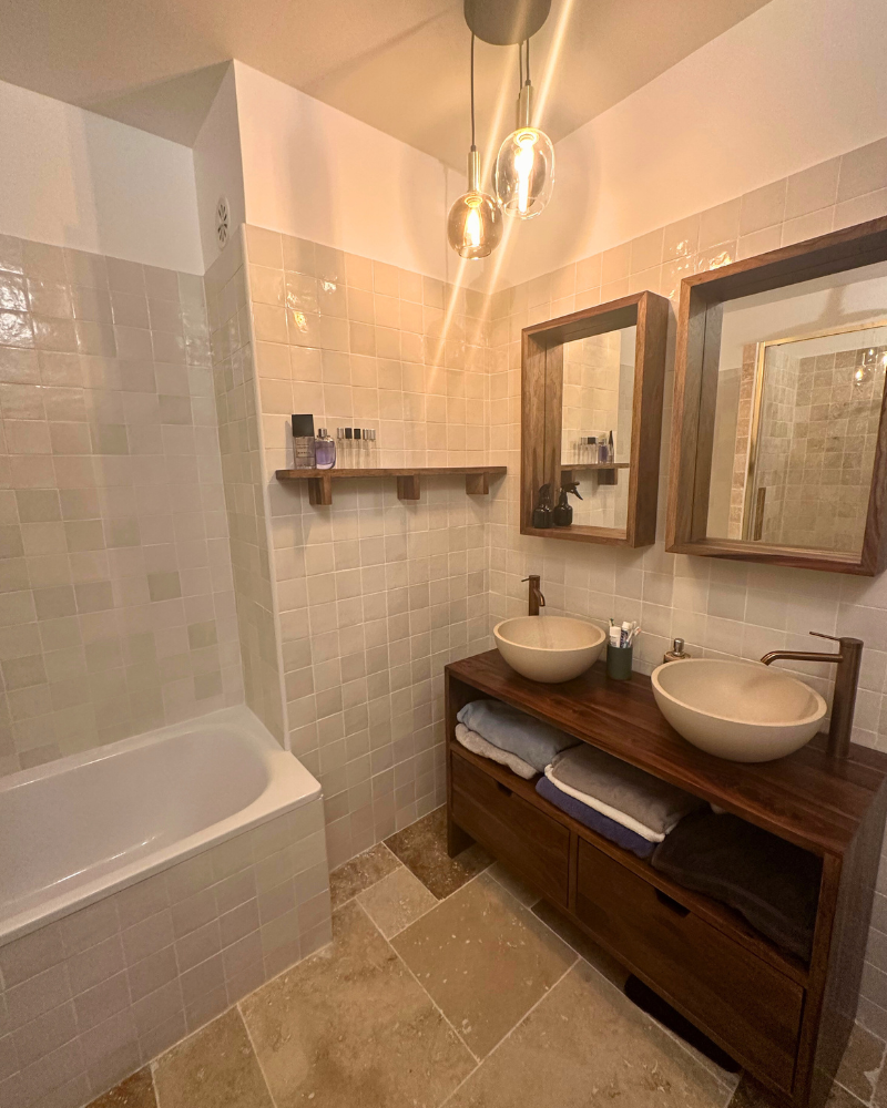 Salle de bains avec baignoire, double vasque sur meuble en bois, miroir, étagère avec produits de toilette, murs et sol carrelés beiges. Plafonnier avec deux ampoules.