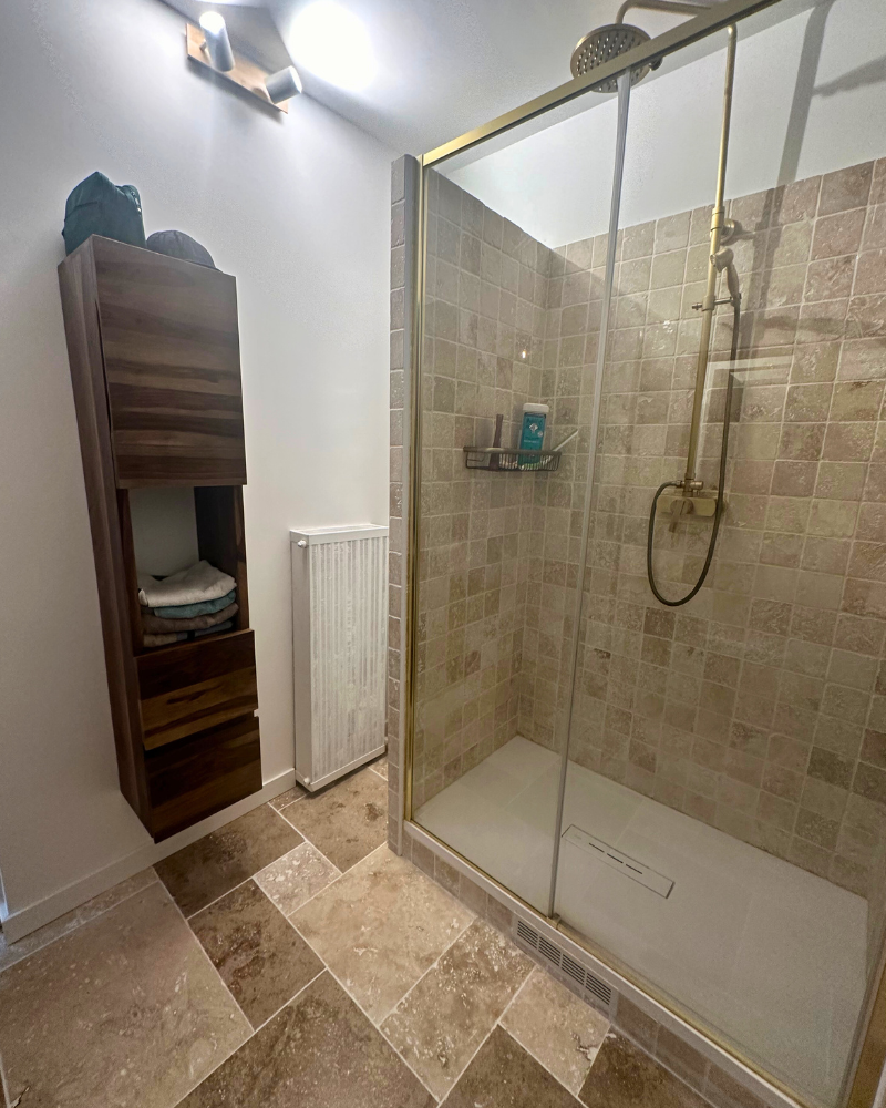 Salle de bain moderne avec douche à l'italienne carrelée, porte vitrée et accessoires dorés. Meuble mural en bois avec serviettes à gauche et radiateur en dessous. Sol et murs en carrelage beige.