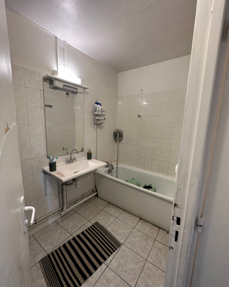 SDB_10_Avant_A Salle de bain avec lavabo, miroir et baignoire. Murs et sol carrelés blancs, tapis rayé au sol et produits de toilette sur une étagère au-dessus de la baignoire.