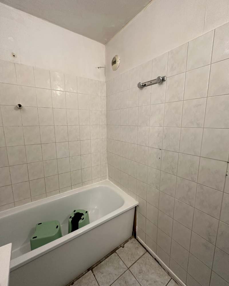 SDB_10_Avant_C Une salle de bain blanche avec un mur carrelé entoure une baignoire avec deux objets verts et noirs à l'intérieur.