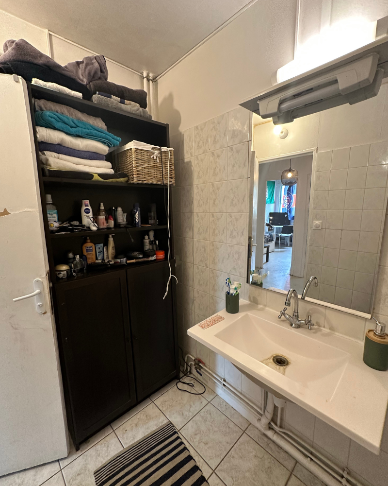 SDB_10_Avant_E Salle de bains avec lavabo, miroir et meuble de rangement avec produits de toilette et serviettes. Un tapis à rayures est posé sur le sol carrelé et une porte mène à une autre pièce.