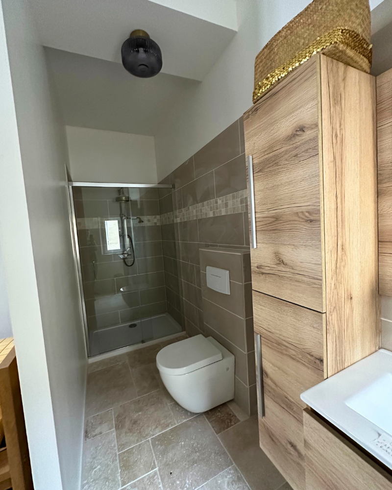 Salle de bains moderne avec toilettes suspendues, douche fermée et meubles en bois. Le sol et les murs sont carrelés et un panier en osier est posé sur le meuble.