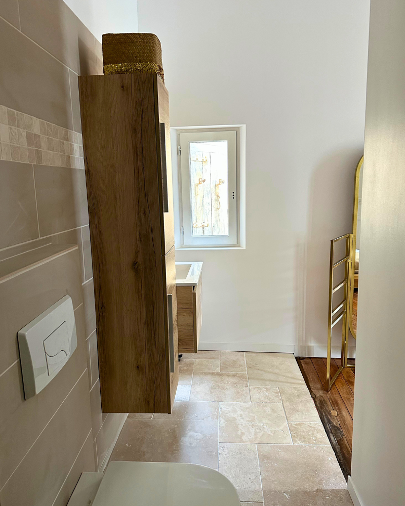 Salle de bain moderne avec carrelage beige, meuble mural en bois, petite fenêtre et miroir à cadre doré. Les toilettes sont partiellement visibles à gauche.