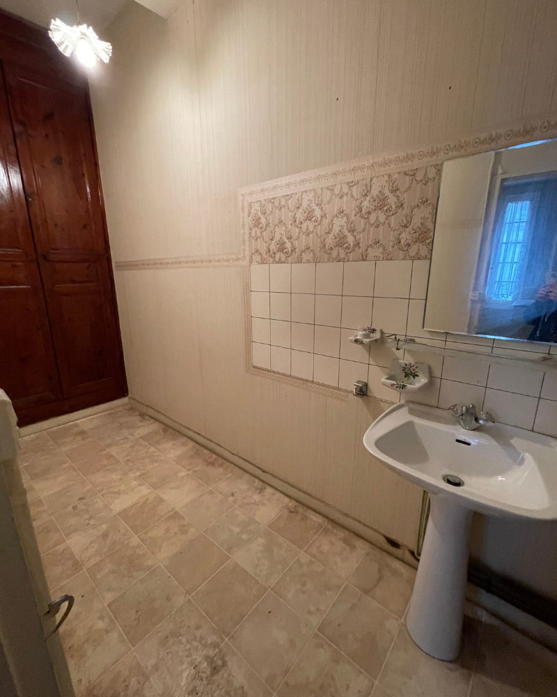 SDB_3_Avant_A Une petite salle de bain avec un lavabo blanc, un miroir, un mur carrelé et des meubles en bois. Le sol est recouvert de carrelage beige.