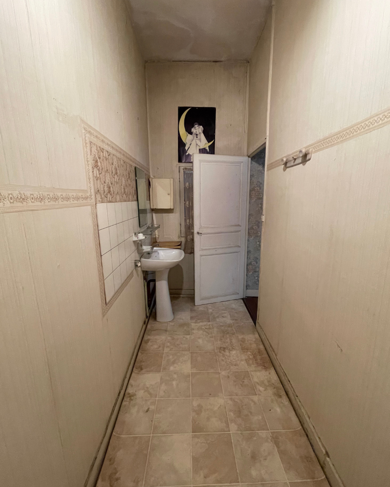 SDB_3_Avant_C Couloir étroit et usé aux murs et au sol carrelés beiges. Un lavabo et un miroir se trouvent à gauche, et une porte blanche fermée se trouve au bout. Une image encadrée se trouve au-dessus de la porte.