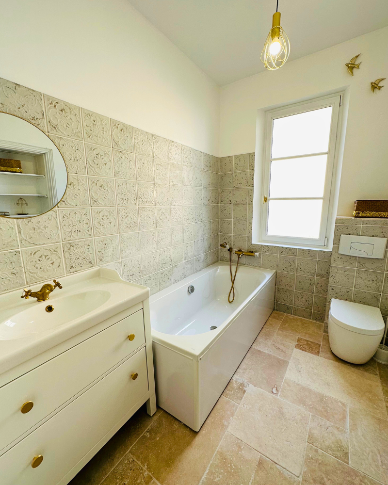Salle de bains avec murs carrelés à motifs, baignoire, accessoires en laiton, meuble-lavabo blanc et suspension. Il y a une fenêtre et des toilettes au design épuré sur un sol carrelé beige.