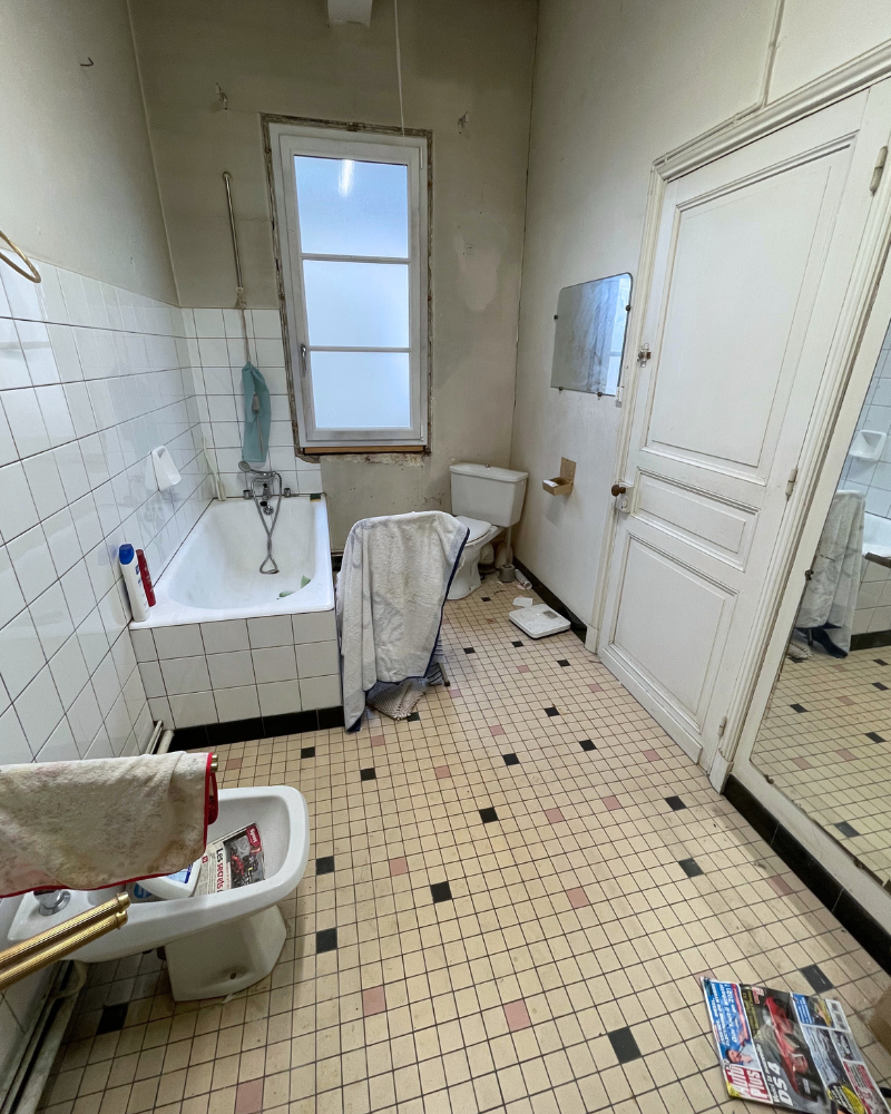 SDB_4_Avant_A Salle de bain ancienne avec carrelage blanc, baignoire, bidet et WC. Une serviette est accrochée près de la baignoire, des magazines sont posés au sol. Une fenêtre au-dessus de la baignoire laisse entrer la lumière. Le miroir semble abîmé.