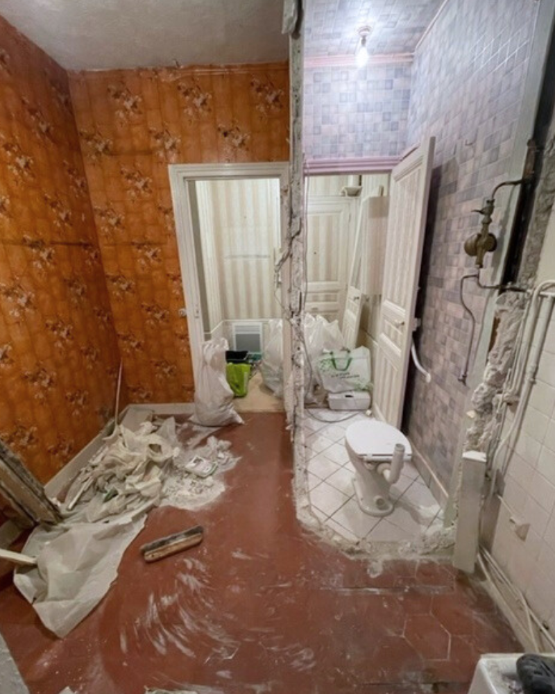 SDB_5_Avant_A Salle de bains partiellement rénovée avec canalisations apparentes, toilettes et matériaux de construction dispersés sur un sol carrelé et béton. Le papier peint fleuri orange a été partiellement retiré des murs.