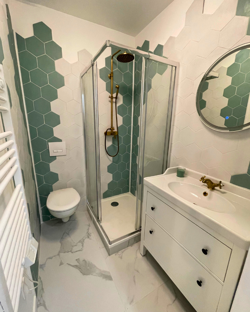 Salle de bain moderne avec douche en verre, carrelage hexagonal vert et blanc, toilettes murales, miroir rond et vanité blanche avec lavabo et deux tiroirs.