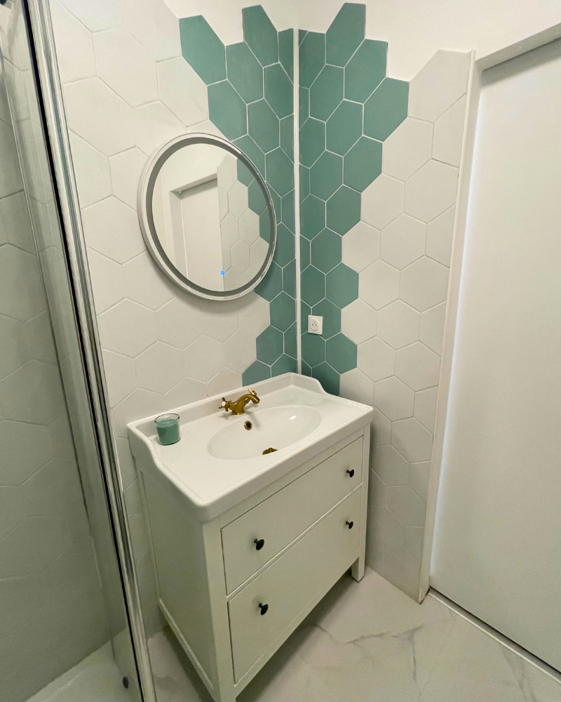 Coin salle de bain avec vanité blanche, robinet doré, carreaux hexagonaux blancs et bleu sarcelle, miroir rond et douche partiellement visible.