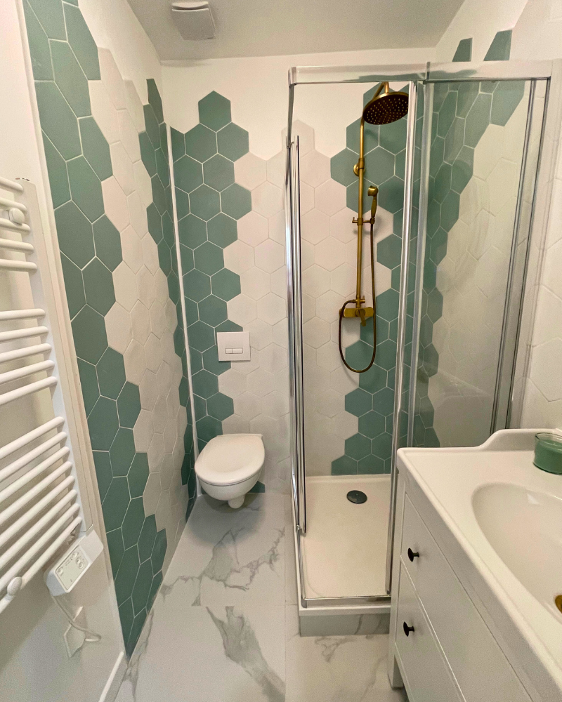 Une petite salle de bain avec des carreaux hexagonaux verts et blancs, des toilettes suspendues, une douche d'angle avec des portes en verre, un lavabo blanc et un sèche-serviettes.