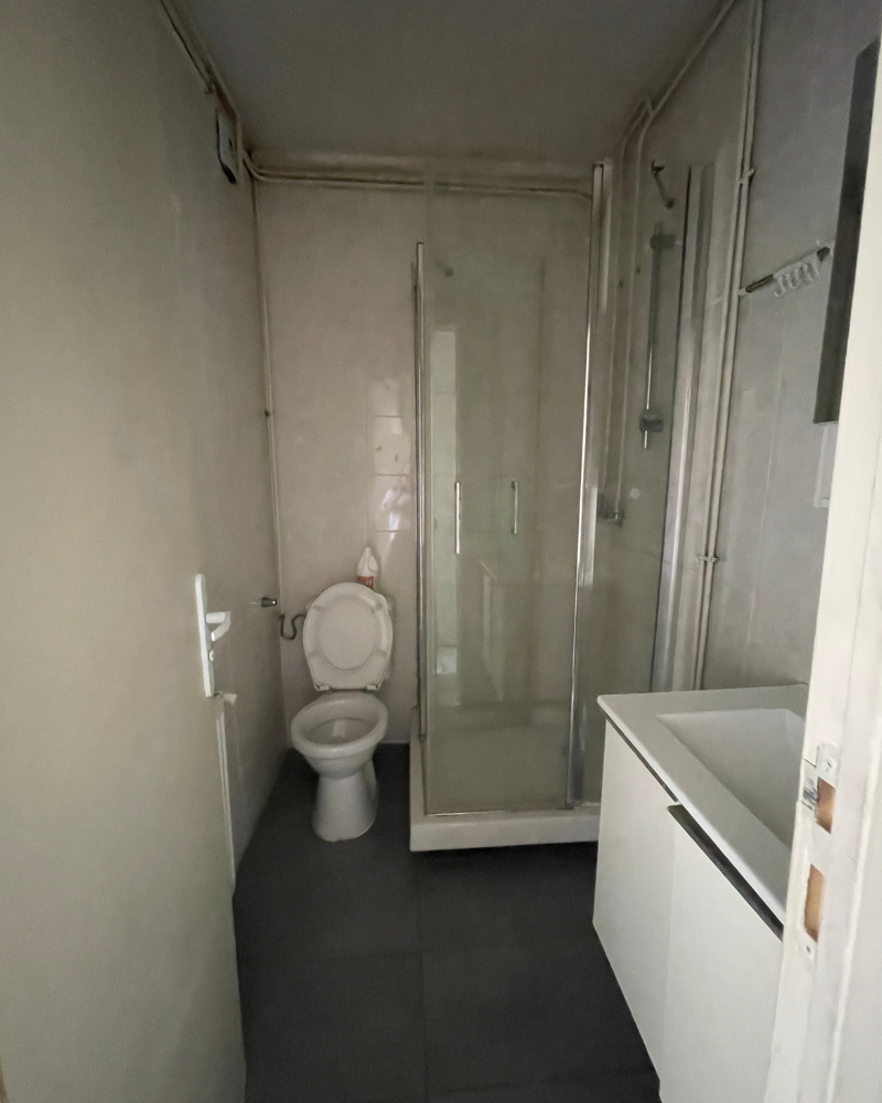 Une salle de bain étroite avec toilettes, douche en verre et lavabo avec meuble. Les murs sont de couleur claire et le sol est gris foncé.