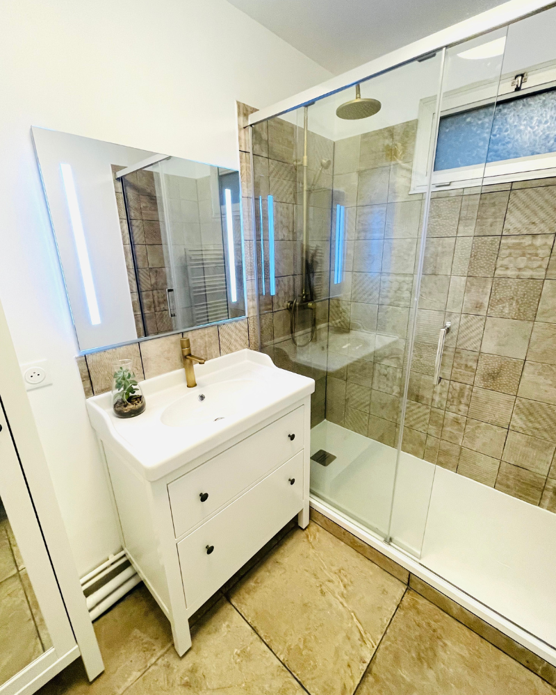 SDB_7_Après_B Salle de bains moderne avec meubles blancs, grand miroir et meuble-lavabo simple. Une plante en pot est posée sur un meuble haut. La porte mène à une douche. Des carreaux clairs recouvrent le sol.