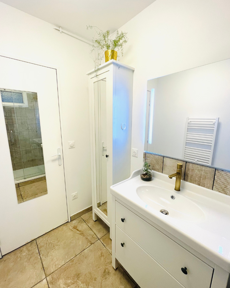 Chambre étroite avec une porte blanche avec un miroir pleine longueur, un sol carrelé, des armoires murales avec des décorations de feuilles et une machine à laver. Un porte-serviettes se trouve sur le mur de gauche.