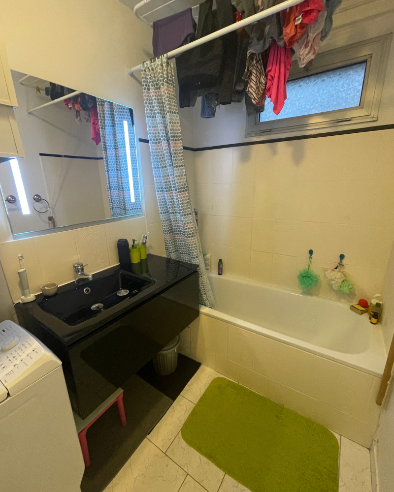 SDB_7_Avant_A (2) Une salle de bain avec un lavabo noir, une petite baignoire, un rideau de douche à motifs et un tapis de bain vert. Des vêtements sont suspendus à une tringle au-dessus et des articles de toilette sont posés sur les bords du lavabo et de la baignoire. Une machine à laver est présente.