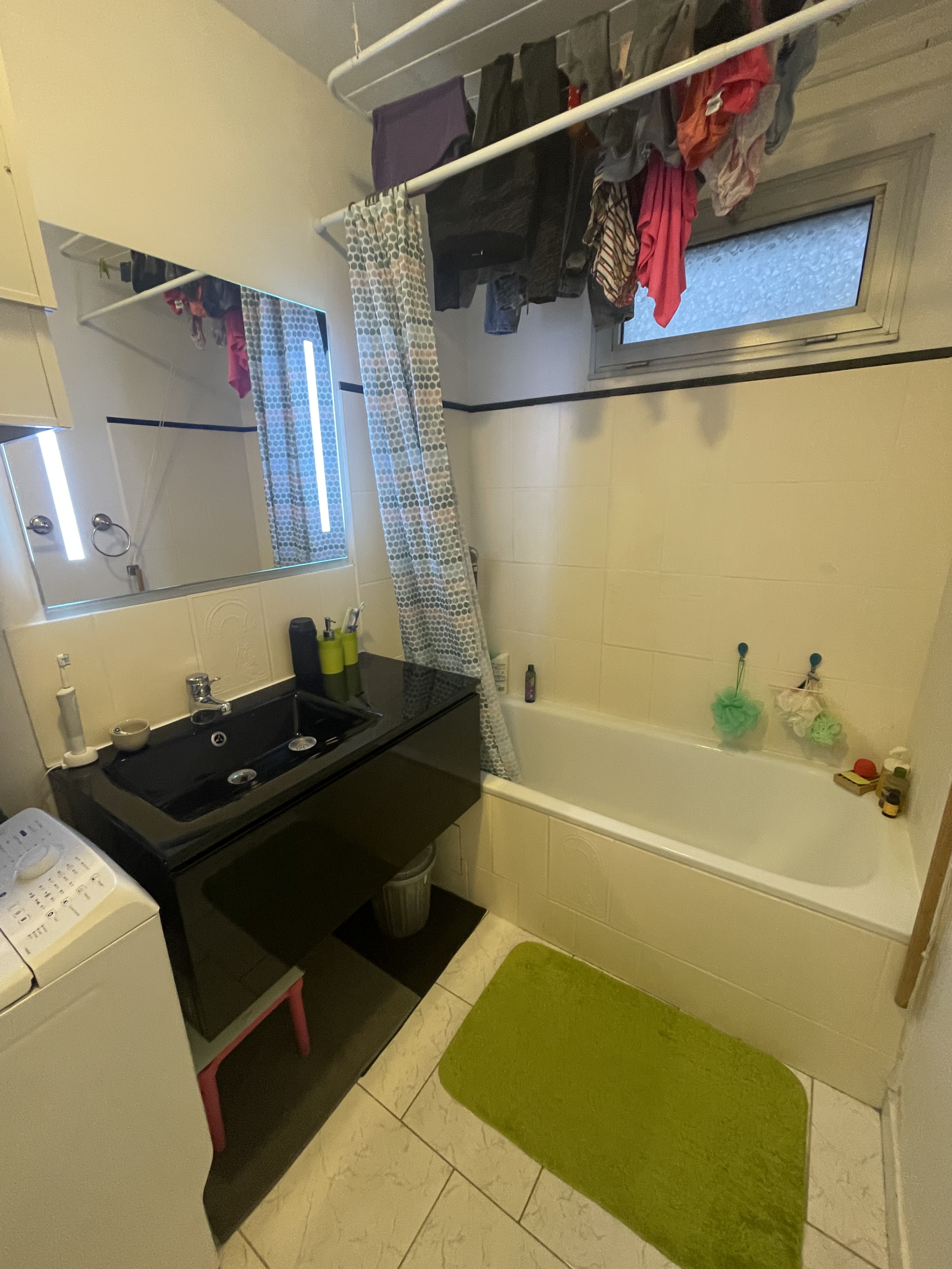 Salle de bain avec lavabo noir, baignoire, tapis vert et vêtements suspendus au-dessus. Une machine à laver se trouve à gauche avec un petit miroir au-dessus du lavabo.
