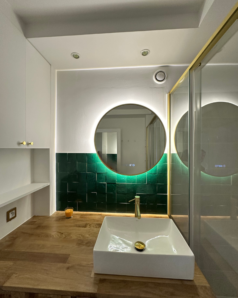 Salle de bain moderne avec miroir rond rétroéclairé, mur carrelé vert et lavabo rectangulaire blanc sur un plan de travail en bois. Une porte de douche en verre se trouve à droite.